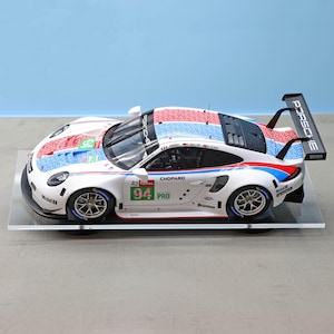 May include: A white Porsche race car model with red, white, and blue accents on the roof and sides. The car has the number 94 and sponsor logos. The rear wing displays "PORSCHE" and "BOSS". The model sits on a clear display stand.
