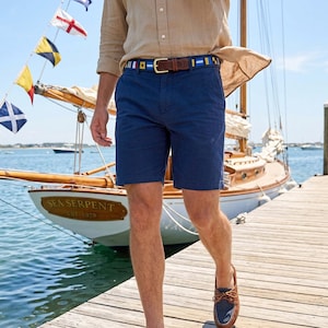 May include: Navy blue shorts with a nautical-themed belt featuring a brown leather strap and colorful flag designs. The person is wearing brown leather boat shoes. A sailboat is in the background.