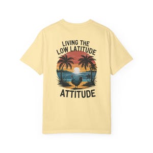 Peut inclure: T-shirt jaune clair avec un motif de coucher de soleil tropical, des palmiers, et un poulet. Le texte dit "LIVING THE LOW LATITUDE ATTITUDE". Le design utilise des couleurs orange, bleu et noir.