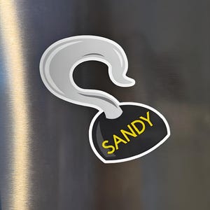 Könnte beinhalten: Ein Magnet im Cartoon-Stil in Form eines Piratenhakens. Der Haken ist silberfarben mit weißem Umriss, und das Etikett darunter ist schwarz mit dem Namen "SANDY" in gelben Buchstaben.