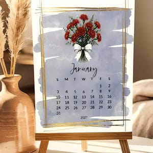 May include: A January 2027 calendar page featuring a watercolor-style blue background with a gold-lined border. The design includes a bouquet of red carnations tied with a white polka-dot bow. The calendar grid displays the days of the month clearly.