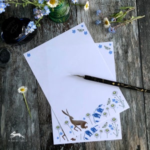Pode incluir: Papel de carta branco com um design floral e de lebre. A papelaria apresenta uma lebre castanha, flores azuis e borboletas. Um frasco de tinta preta e uma caneta estão sobre uma superfície de madeira rústica, com margaridas e miosótis.