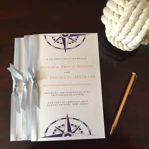 May include: A white wedding invitation with a compass design and gold accents. The invitation reads "A Celebration of Marriage Jessica Dawn Simms and Ian Michael O'Grady Saturday, the thirtieth of April Two Thousand Sixteen Our Lady of Perpetual Help Seaside Heights, New Jersey".