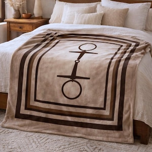 May include: A tan and brown blanket with a horse bit design. The blanket features a large, dark brown horse bit graphic in the center, framed by a series of brown stripes. The blanket is draped over a bed with white bedding.