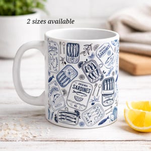 Könnte beinhalten: Weiße Keramik-Tasse mit einem sich wiederholenden Design von blau-weißen Sardinenbüchsen. Die Tasse zeigt verschiedene Sardinenbüchsen-Illustrationen, Olivenzweige und den Text "Original Sardines". Der Text "2 Größen verfügbar" ist ebenfalls sichtbar.