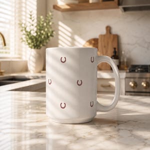 May include: A white ceramic mug featuring a repeating pattern of small, dark red horseshoe icons distributed across its exterior surface. The mug has a smooth, cylindrical body and a curved handle on the right side, designed for holding hot beverages.