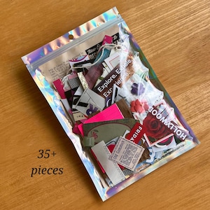 May include: A holographic resealable plastic pouch containing over 35 assorted paper ephemera pieces, including vintage-style text snippets, floral imagery, and a red rose graphic with the word Hollywood, all visible through the clear material.