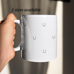 Könnte beinhalten: Eine weiße Keramiktasse mit einem Hufeisenmuster. Die Tasse hat einen Henkel und der Text "2 Größen verfügbar" ist oben aufgedruckt.