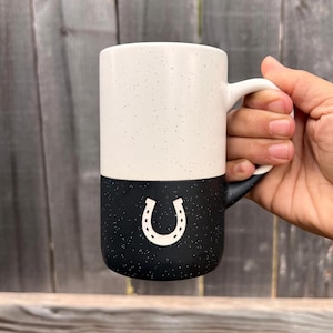 May include: A two-toned ceramic mug featuring a speckled cream upper section and a speckled black lower section. The black portion displays a white horseshoe icon centered on the front. The mug has a curved handle on the right side.