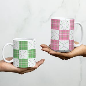 Könnte beinhalten: Zwei weiße Keramikbecher mit einem karierten Muster in Grün und Rosa. Die Becher haben ein sich wiederholendes Muster aus grünen und rosa Quadraten mit kleinen weißen und grünen geometrischen Formen. Die Becher werden in Händen gehalten.