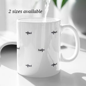 May include: White ceramic mug with a handle, decorated with small, dark gray shark silhouettes. The text "2 sizes available" is printed near the top. Water is being poured into the mug.