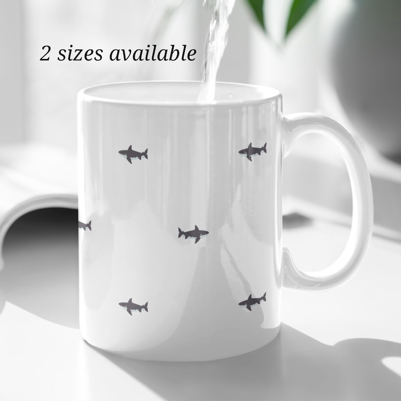 May include: White ceramic mug with a handle, decorated with small, dark gray shark silhouettes. The text "2 sizes available" is printed near the top. Water is being poured into the mug.