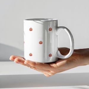 May include: White ceramic mug decorated with small orange pumpkin illustrations. The mug has a large handle and is designed for hot beverages. A festive and seasonal item, perfect for Halloween or autumn.