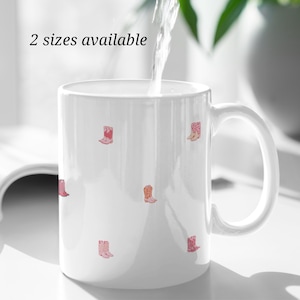 May include: White ceramic mug decorated with small pink and orange cowboy boot illustrations. The text "2 sizes available" is printed on the top left of the mug. Water is being poured into the mug.