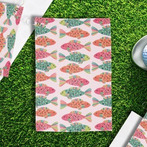 May include: Golf towel with a repeating pattern of colorful fish in shades of pink, orange, green, and blue. The fish have polka dots and striped fins. The towel is on a green surface.