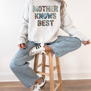 May include: A light gray sweatshirt with the text "MOTHER KNOWS BEST" in a floral pattern. The sweatshirt is paired with light blue jeans and white sneakers with black stripes. The person is sitting on a wooden stool.
