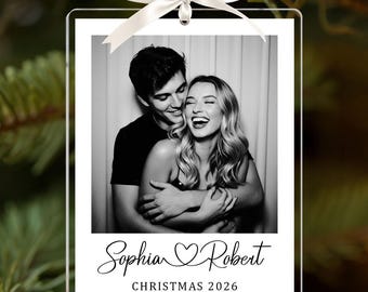 May include: A rectangular clear acrylic ornament featuring a black and white photograph of a couple. The bottom section displays the names Sophia and Robert with a heart symbol in between, followed by the text CHRISTMAS 2026. A white ribbon bow is attached.