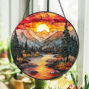 May include: A round stained glass window hanging with a chain. The window depicts a sunset over a lake and mountains with a bright orange sun and a dark blue sky.
