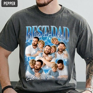May include: Dark gray t-shirt with a graphic that says "BEST DAD" in blue, with a collage of photos. The photos show a group of men and children. The text "Est. 2018" is also on the shirt.