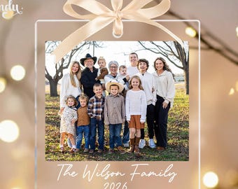May include: A square clear acrylic ornament featuring a printed photograph of a large family standing outdoors. The ornament is topped with a beige satin ribbon bow and includes the white printed text The Wilson Family and the year 2026.