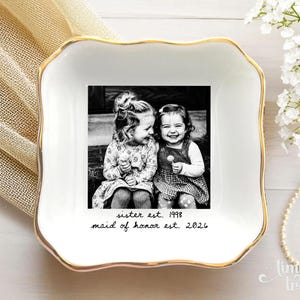 May include: A square white ceramic trinket dish with a gold-trimmed scalloped edge. The center features a black and white photograph of two young children, with the text "sister est. 1998" and "maid of honor est. 2026" printed below.