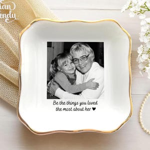 May include: A square white ceramic trinket dish with a gold-trimmed scalloped edge. The center features a black and white photograph of a child hugging an adult, with the printed text: Be the things you loved the most about her ♥.
