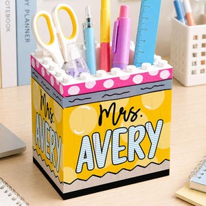 May include: A square pencil holder constructed from yellow, white, and pink building blocks. The front features the text "Mrs. AVERY" in black and blue lettering. It contains white scissors, a blue pen, a yellow marker, a pink pen, and a blue ruler.