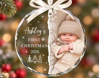 May include: A round glass Christmas ornament featuring a white ribbon bow at the top. The left side displays the text "Ashley's FIRST CHRISTMAS 2026" with snowflake and tree icons. The right side shows a photograph of a baby in a knit hat.