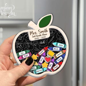 May include: Apple-shaped wooden magnet with a chalkboard design, featuring the text "Mrs. Smith 3rd Grade Class 2025-2026" and student names on crayon-shaped labels. The magnet is held by a hand.