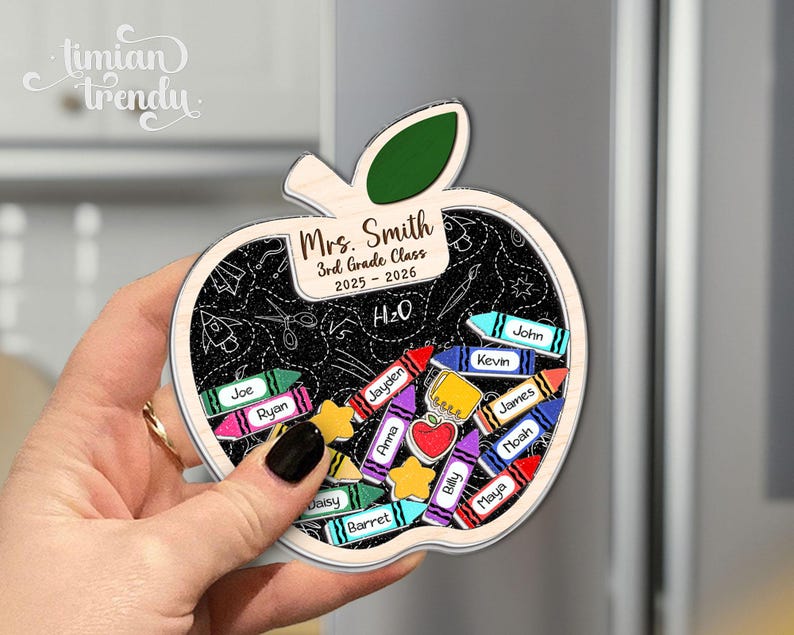 May include: Apple-shaped wooden magnet with a chalkboard design, featuring the text "Mrs. Smith 3rd Grade Class 2025-2026" and student names on crayon-shaped labels. The magnet is held by a hand.