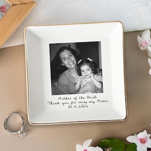 May include: A square white ceramic trinket dish with a gold-toned rim. The center features a black and white photograph of an adult and a child, with the text "Mother of the Bride, Thank you for being my Mama, 06.15.2026" printed below it.