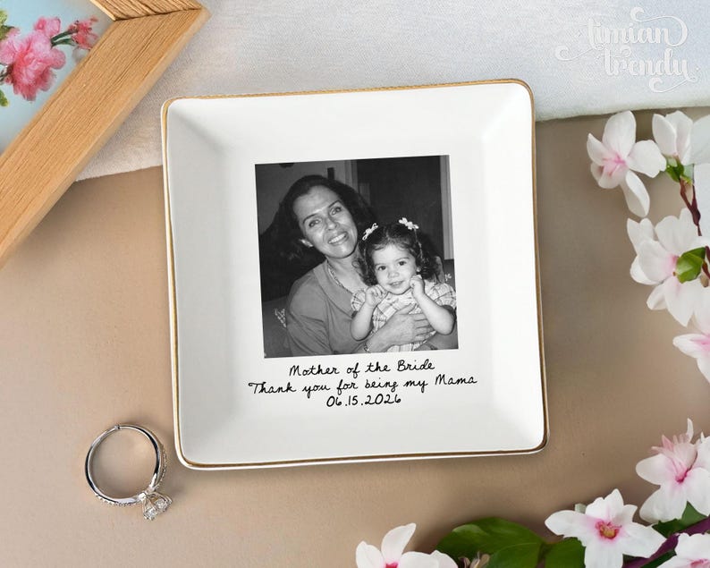May include: A square white ceramic trinket dish with a gold-toned rim. The center features a black and white photograph of an adult and a child, with the text "Mother of the Bride, Thank you for being my Mama, 06.15.2026" printed below it.