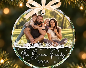 May include: A round, clear acrylic Christmas ornament featuring a photograph of a family of three sitting outdoors. The ornament is topped with a cream-colored ribbon bow and displays the text The Brown Family and 2026 in white script.