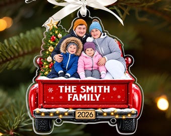 May include: A custom Christmas ornament featuring a family of four sitting in the back of a red pickup truck. The truck bed displays the text THE SMITH FAMILY and the year 2026. A white ribbon is attached to the top for hanging.