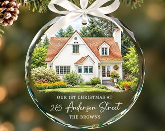 May include: A round, clear glass Christmas ornament featuring a white house with a red roof, green trees, and colorful flowers. The text reads: OUR 1ST CHRISTMAS AT 265 Anderson Street THE BROWNS. A white satin ribbon is tied at the top.
