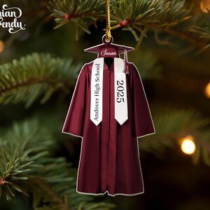 May include: A maroon graduation gown ornament with a white sash that reads "Andover High School 2025." The ornament includes a graduation cap with the name "Susan." The ornament is hanging from a gold string against a blurred background of a Christmas tree.