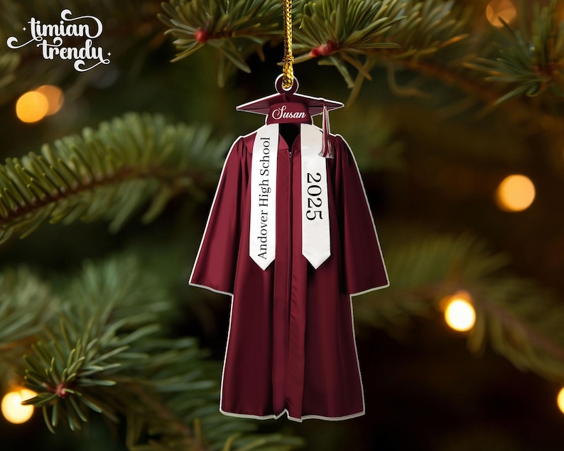 May include: A maroon graduation gown ornament with a white sash that reads "Andover High School 2025." The ornament includes a graduation cap with the name "Susan." The ornament is hanging from a gold string against a blurred background of a Christmas tree.