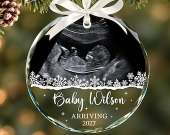 May include: A round, clear glass Christmas ornament featuring a black and white ultrasound image of a fetus. The ornament is decorated with white snowflakes and the text Baby Wilson ARRIVING 2027, finished with a white satin ribbon bow.