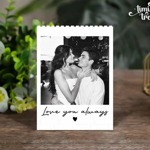 Puede incluir: Un bloque rectangular blanco con un borde de patr&oacute;n de ladrillo que presenta una fotograf&iacute;a en blanco y negro de una pareja abraz&aacute;ndose. Debajo de la foto, el texto Love you always aparece en una fuente cursiva negra, seguido de un coraz&oacute;n.