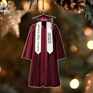 May include: A burgundy graduation gown ornament with a white sash that reads "Andover High School 2025." The ornament includes a graduation cap with the name "Susan." The ornament is hanging from a gold string.