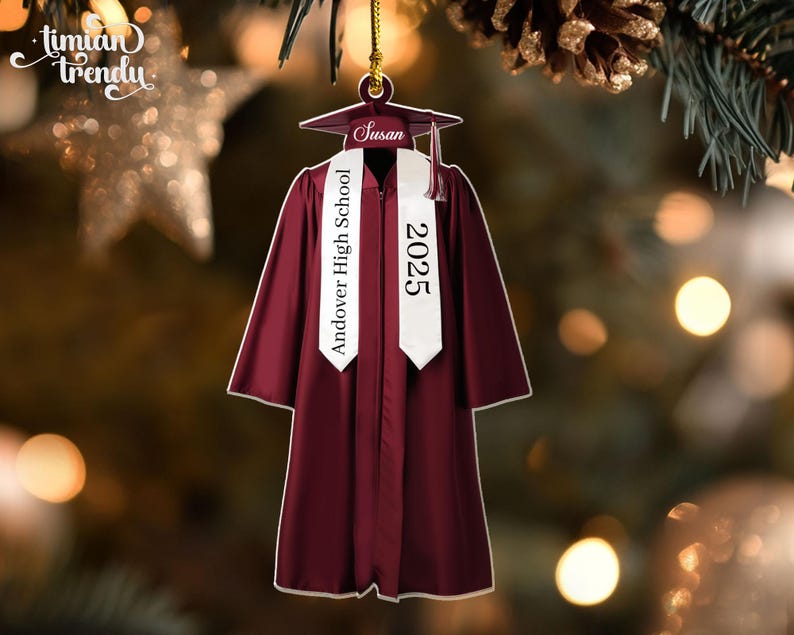May include: A burgundy graduation gown ornament with a white sash that reads "Andover High School 2025." The ornament includes a graduation cap with the name "Susan." The ornament is hanging from a gold string.