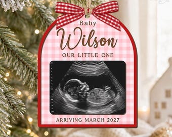 May include: An arched Christmas ornament featuring a pink and white gingham pattern, a red and white checkered bow, and a central ultrasound image. Text reads Baby Wilson, OUR LITTLE ONE, and ARRIVING MARCH 2027 in brown and black lettering.