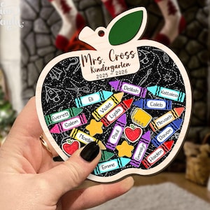 May include: Wooden apple-shaped ornament with a chalkboard design. It features the text "Mrs. Cross Kindergarten 2025-2026" and crayon illustrations with student names. A green leaf tops the apple.