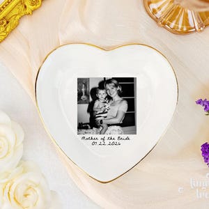 May include: A white heart-shaped ceramic trinket dish with a gold-painted rim. The center features a black and white photograph of a person holding a child, with the text "Mother of the Bride" and the date "07.22.2026" printed in black below it.