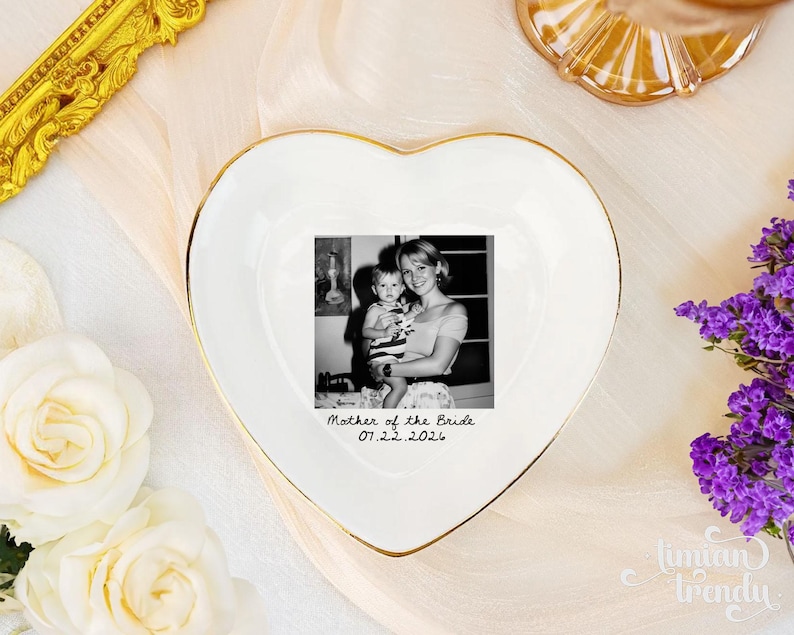 May include: A white heart-shaped ceramic trinket dish with a gold-painted rim. The center features a black and white photograph of a person holding a child, with the text "Mother of the Bride" and the date "07.22.2026" printed in black below it.