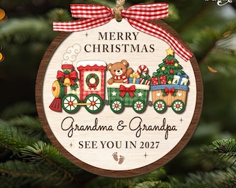 May include: A round wooden Christmas ornament featuring a red and green train illustration with a teddy bear, a Christmas tree, and gifts. The text reads MERRY CHRISTMAS, Grandma & Grandpa, and SEE YOU IN 2027, with a red and white gingham bow.