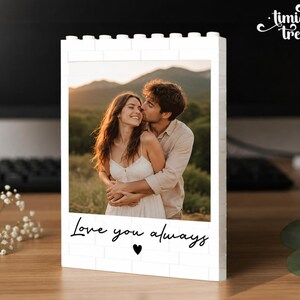 Puede incluir: Un marco de fotos rectangular blanco construido con bloques de construcci&oacute;n entrelazados. El marco muestra una fotograf&iacute;a de una pareja abraz&aacute;ndose al aire libre. Debajo, el texto "Love you always" est&aacute; impreso en negro con un coraz&oacute;n.