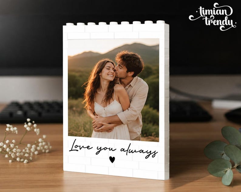 Puede incluir: Un marco de fotos rectangular blanco construido con bloques de construcci&oacute;n entrelazados. El marco muestra una fotograf&iacute;a de una pareja abraz&aacute;ndose al aire libre. Debajo, el texto "Love you always" est&aacute; impreso en negro con un coraz&oacute;n.