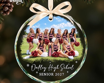 May include: A round, clear glass ornament featuring a photograph of a cheerleading squad in black and red uniforms with white pom-poms on a field. The ornament is topped with a cream-colored ribbon bow and includes the text Oakley High School SENIOR 2027.