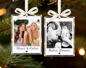 May include: Two rectangular clear acrylic photo ornaments hanging from white satin ribbons. The left ornament features a color photo of two people blowing bubbles with the text Olivia & Katie and a heart. The right ornament shows a black and white photo of two people with the text Bestie Forever and a heart.